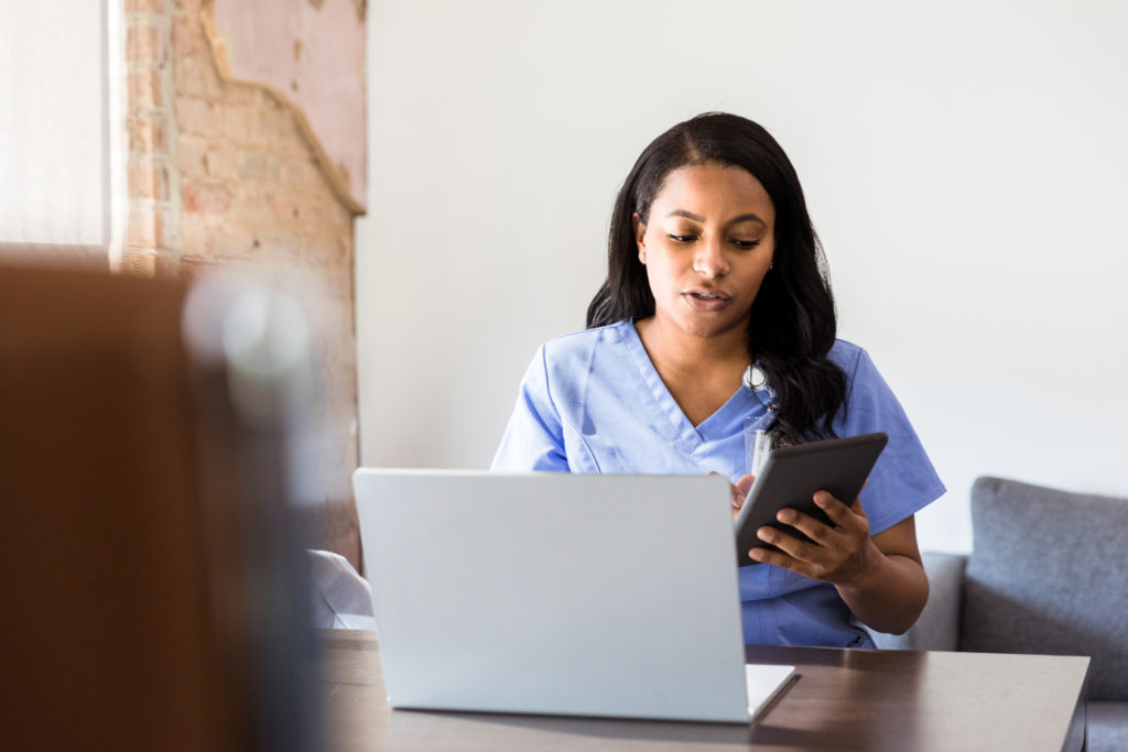 A serious doctor checks her appointment calendar while working in her office. She is using a digital tablet to check her calendar. She is sitting in front of an open laptop.