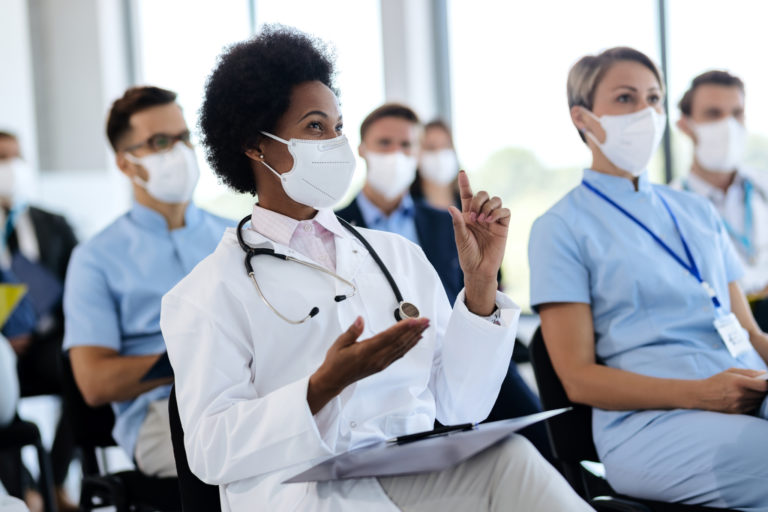 Happy African American female doctor with protective face mask asking a question while attending a seminar in board room.