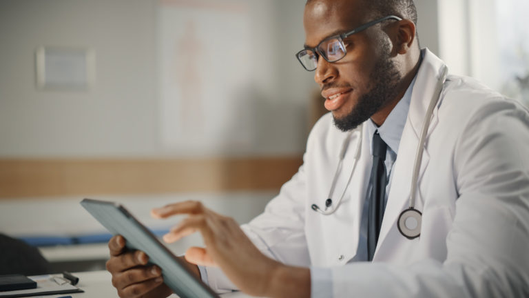 Happy and Smiling African American Male Doctor Wearing White Coat Working on Tablet Computer at His Office. Medical Health Care Professional Working with Test Results, Patient Treatment Planning.