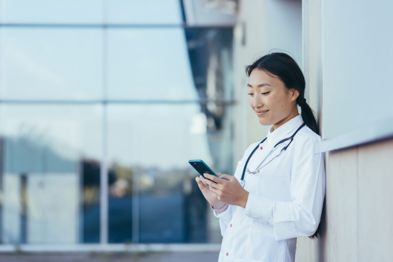 Happy female doctor, smiling and rejoicing, Asian woman uses the phone, during a break near the clinic medical apps, looking at screen, working online, chatting, consulting patient, telemedicine concept