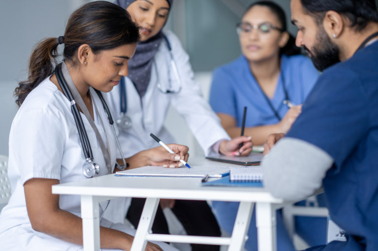 A small group of four nurses sit together around a table as they meet to discus patient cases and their plans of care. They are each dressed professionally in scrubs and have stethoscopes around their necks and various files and papers can be seen spread out on the table in front of them. They are each looking down at the documents on the table as a female nurse shares them with the group.